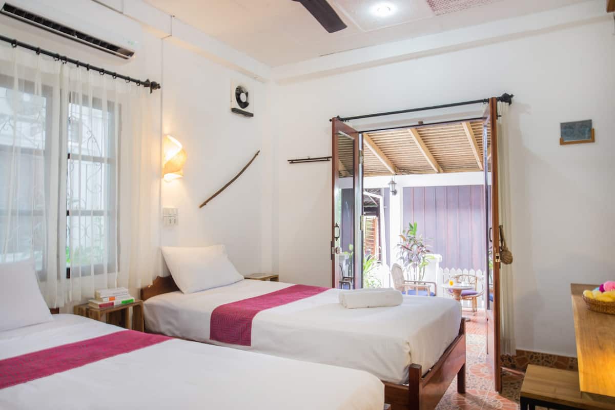 A hotel room with two beds, a window with curtains, a wall-mounted fan, and a sliding door leading to an outdoor area with a chair and table.