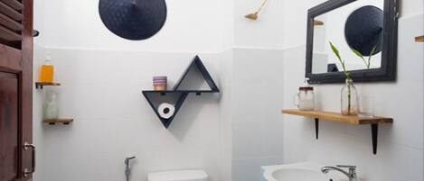 Baño con inodoro, lavabo y espejo. Se observa un estante con artículos de tocador y un objeto decorativo en la pared.