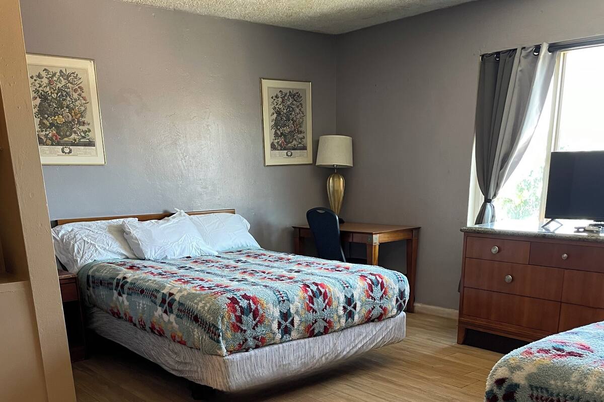 A hotel room with a bed, a desk, a chair, a dresser, and two framed pictures on the wall.