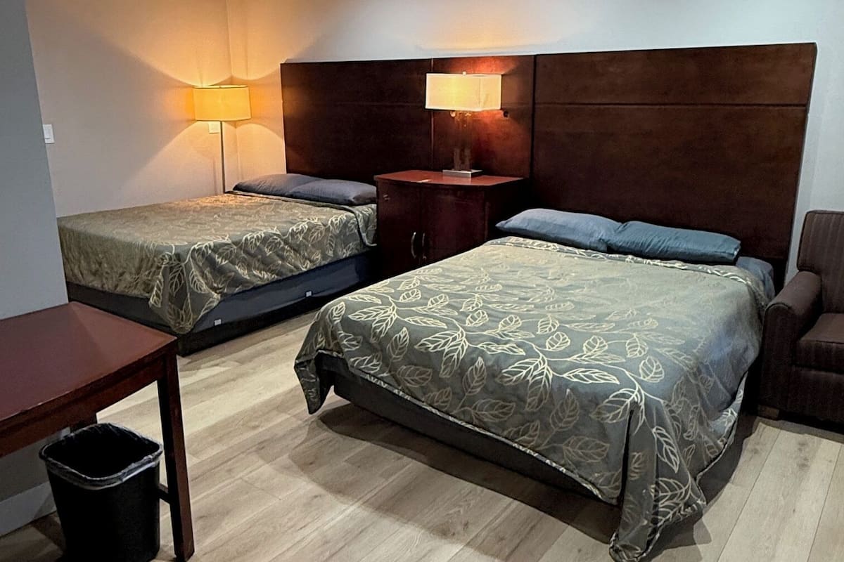 A hotel room with two beds, a wooden table, a trash bin, and a nightstand with a lamp.