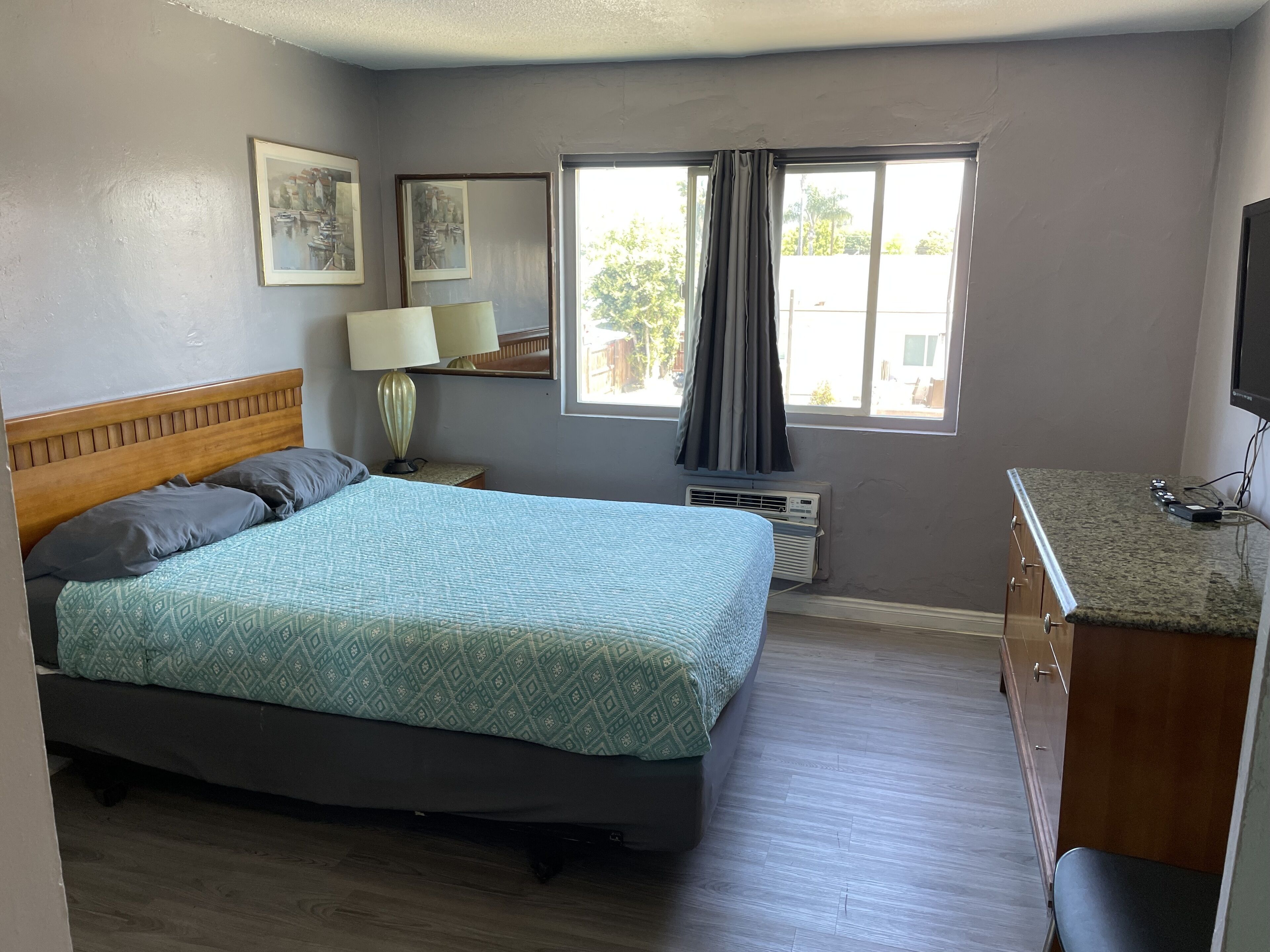 A bedroom with a bed, a nightstand, a lamp, a window with curtains, a television, and a dresser.
