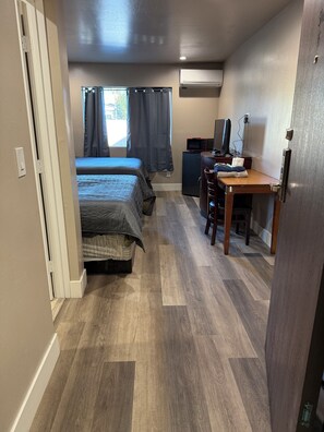 Habitación de hotel compacta con dos camas, un escritorio con ordenador, un refrigerador pequeño y un equipo de aire acondicionado.
