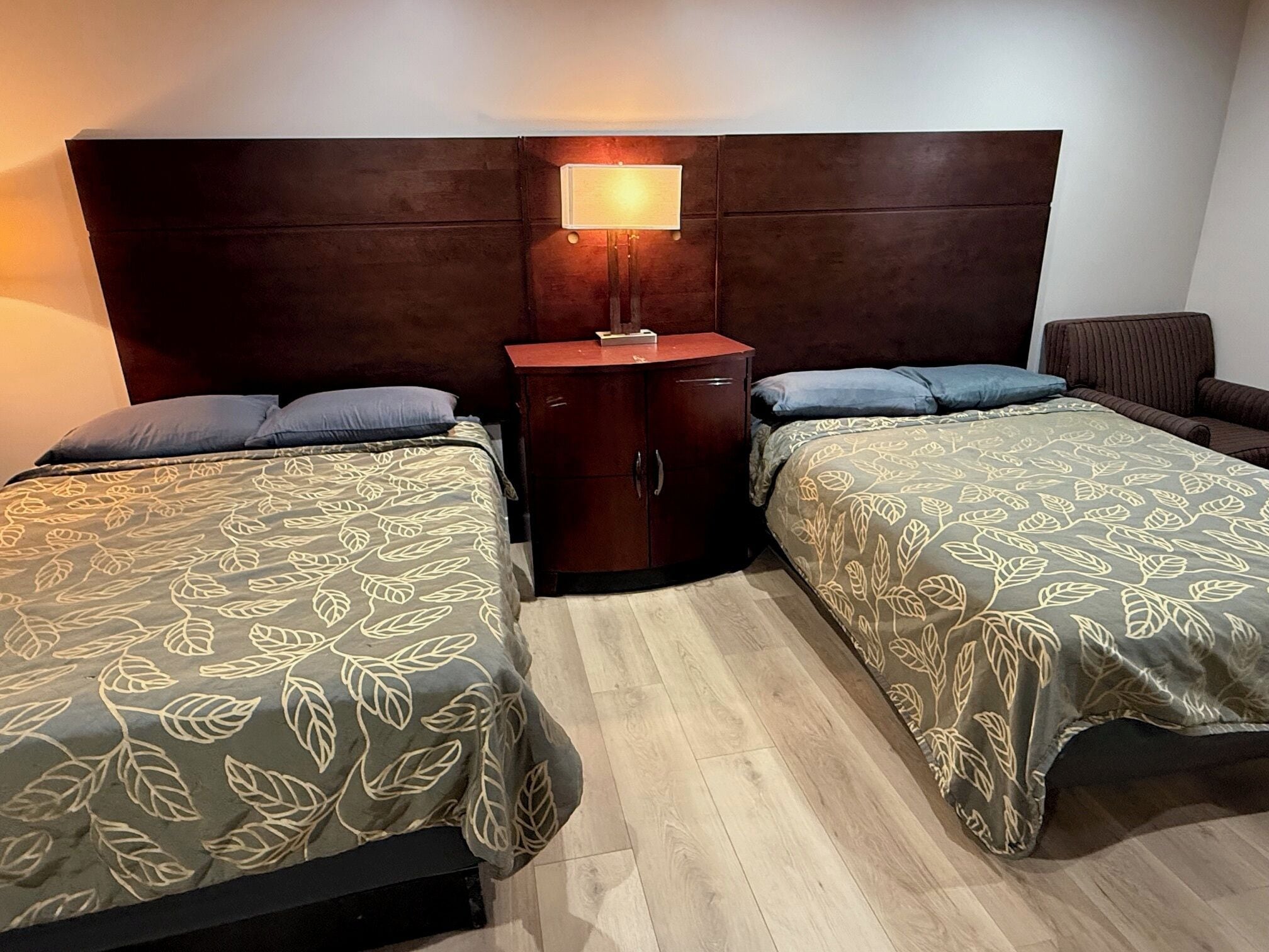 A hotel room with two beds, a nightstand, a lamp, and a chair.