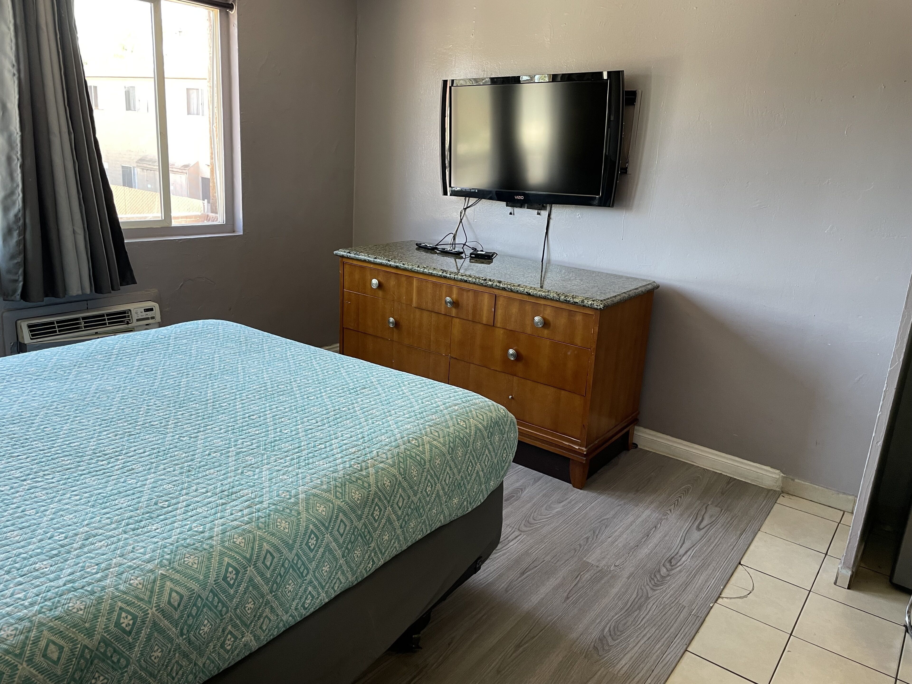Un dormitorio con cama, cómoda, televisor montado en la pared y ventana con cortinas.