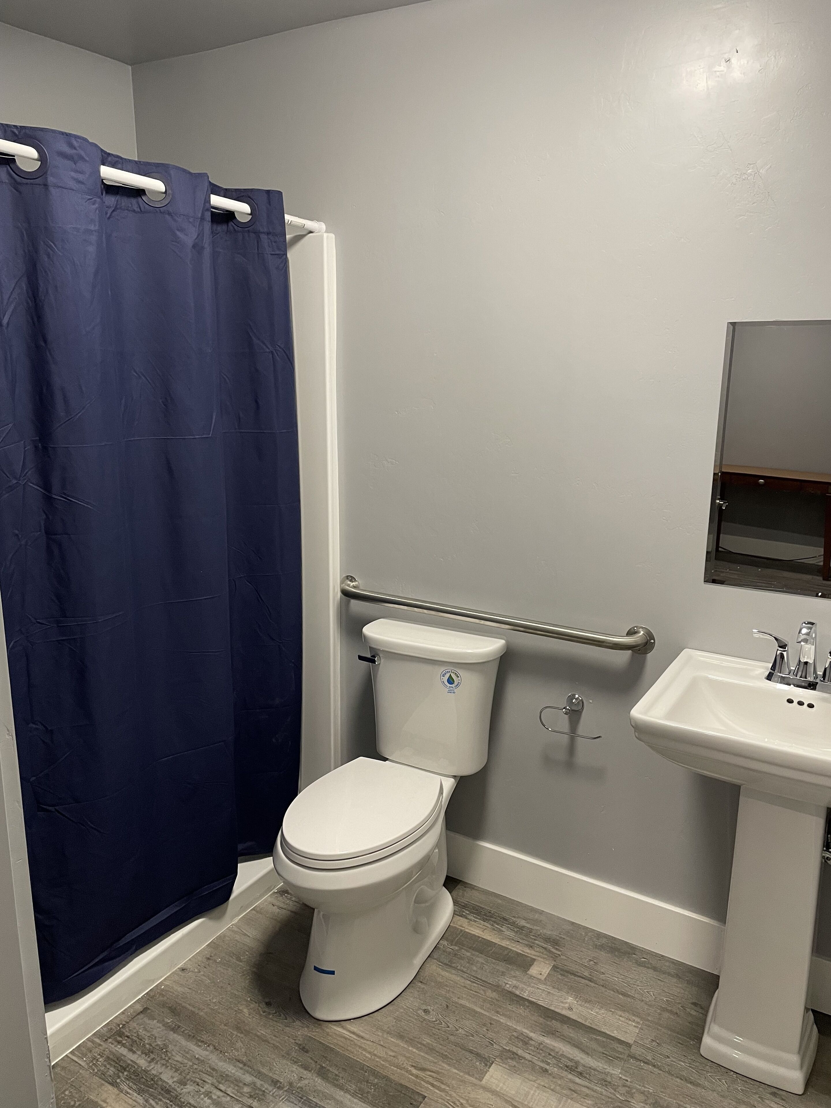Un baño con un inodoro blanco, una cortina de ducha azul, un lavabo blanco y suelo de madera.