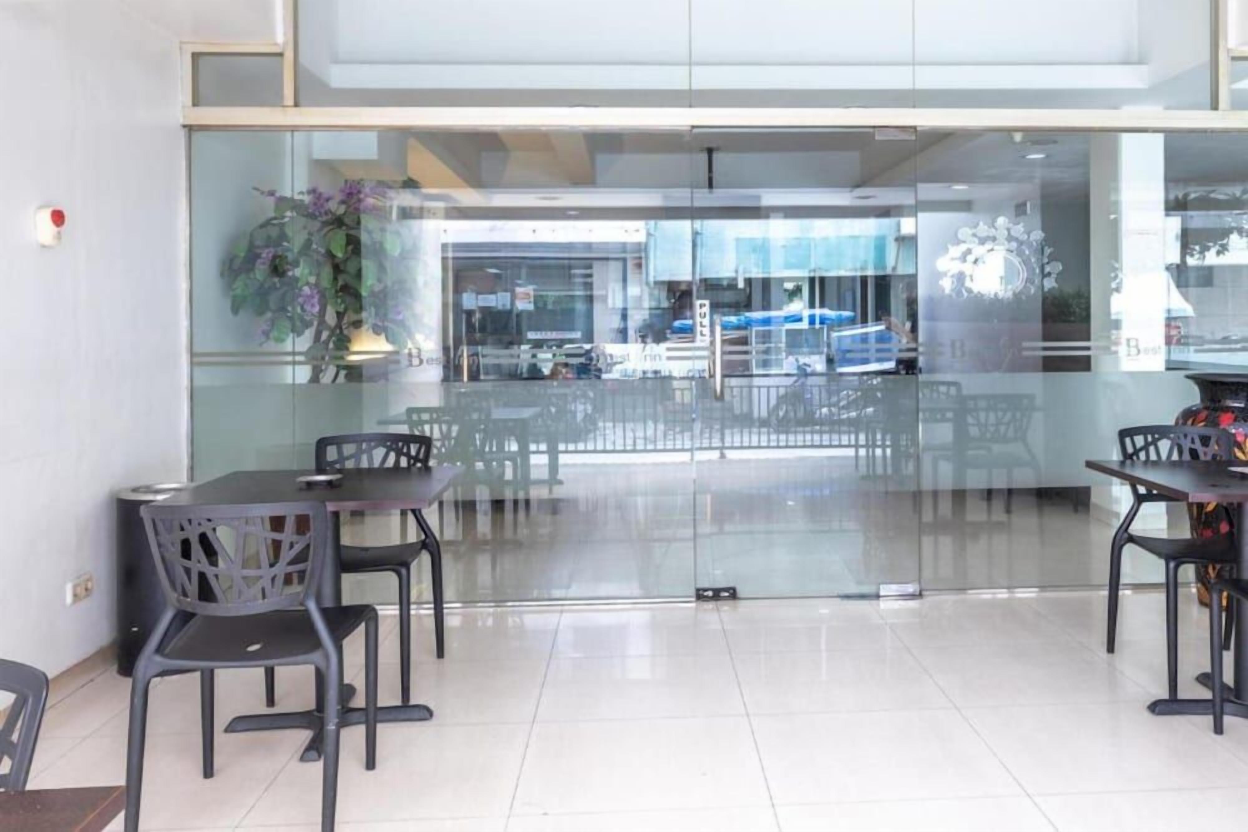 A modern interior with a glass partition, several tables and chairs, and a visible outdoor area with cars.