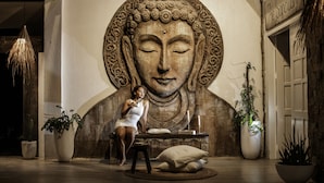 A large Buddha mural in a lobby with a woman sitting on a bench. There are potted plants and a hanging light fixture. - Buena Vida Resort and Spa (Daanbantayan)