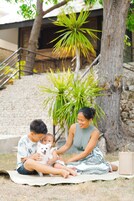 Una familia sentada sobre una esterilla al aire libre, con un perro, árboles y un edificio al fondo.