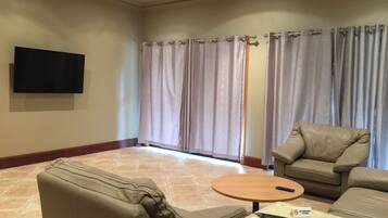 A living room with a flat-screen TV mounted on the wall, a beige sofa, a round coffee table, and a 'No Smoking' sign on the floor.