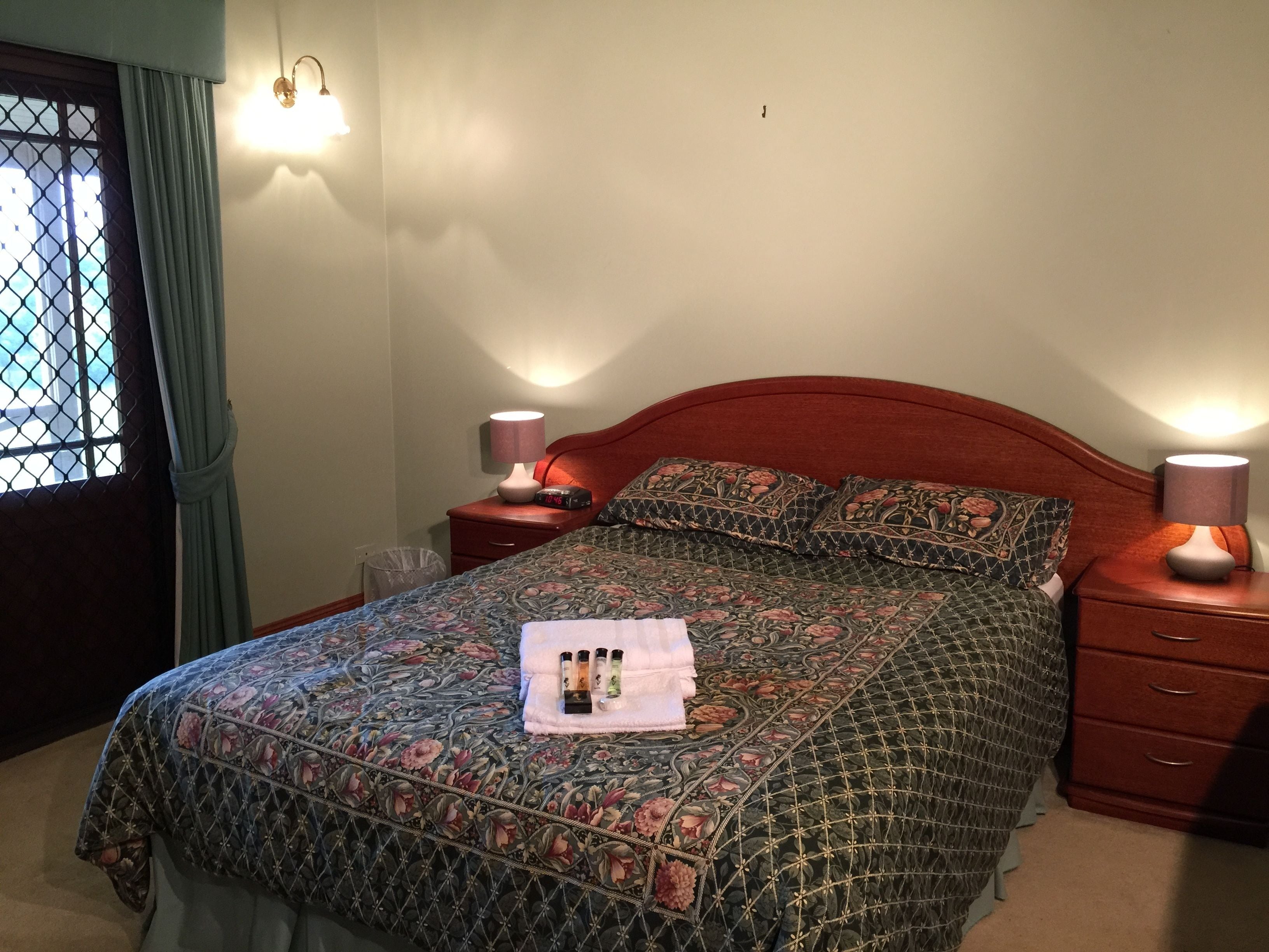 A neatly made bed with floral bedding, two bedside lamps, a nightstand with a clock, and a window with curtains.