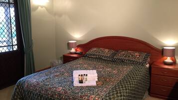 A neatly made bed with floral bedding, two bedside lamps, a nightstand with a clock, and a window with curtains.