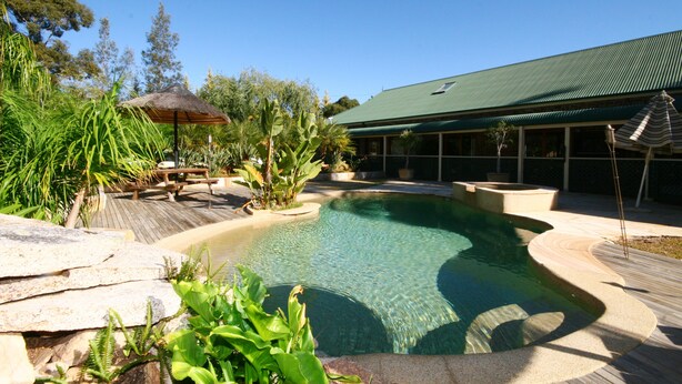 A swimming pool with a curved edge, surrounded by a wooden deck and lush greenery. - Relaxin (Middle Dural)