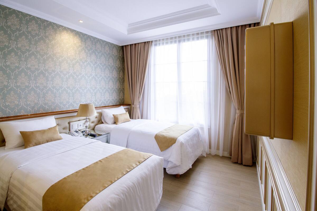Ein Hotelzimmer mit zwei Betten, einem Nachttisch, einer Lampe und einem großen Fenster mit Vorhängen.