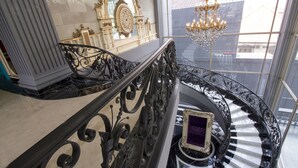 A grand staircase with ornate black railings and a chandelier, leading to a room with a checkered floor and a purple framed artwork. - Park View Hotel (Bandung)