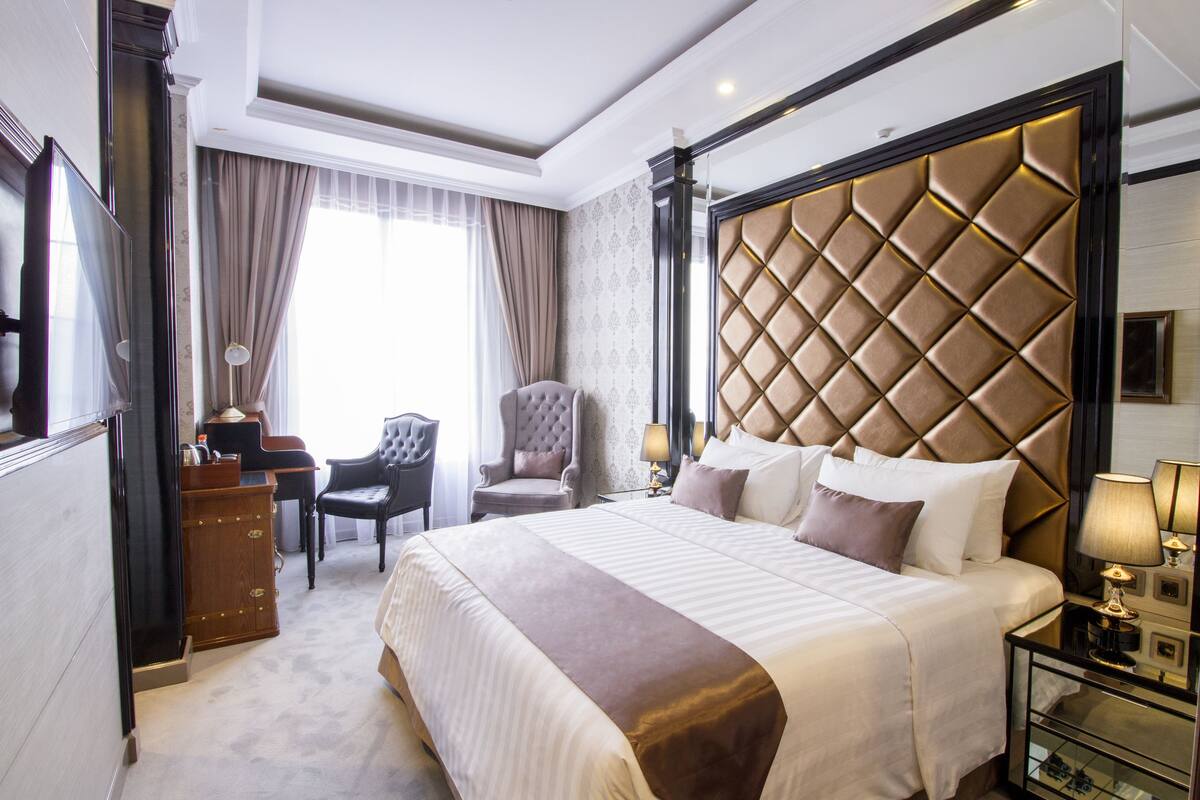 Ein Hotelzimmer mit einem großen Bett, einem hölzernen Schminktisch, einem Sessel und einem Schreibtisch mit Lampe.