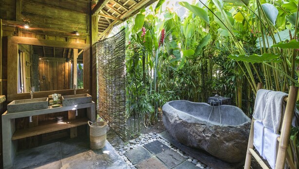 A rustic bathroom with a stone sink, a large stone bathtub, and lush greenery surrounding the space.