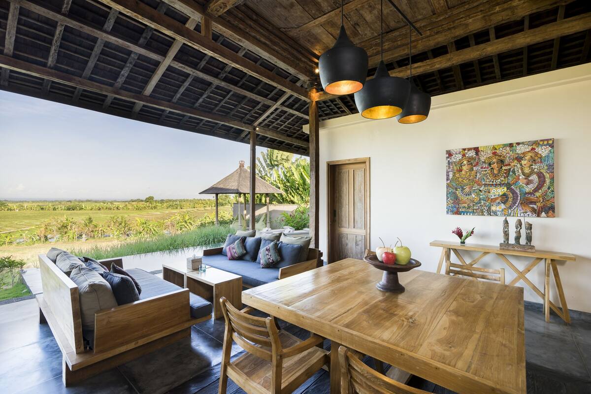 A spacious room with a wooden dining table, a sofa with cushions, and a view of a lush green field.