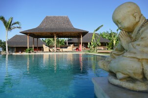Una piscina con una estatua, una zona de estar cubierta y un edificio con tejado de paja.