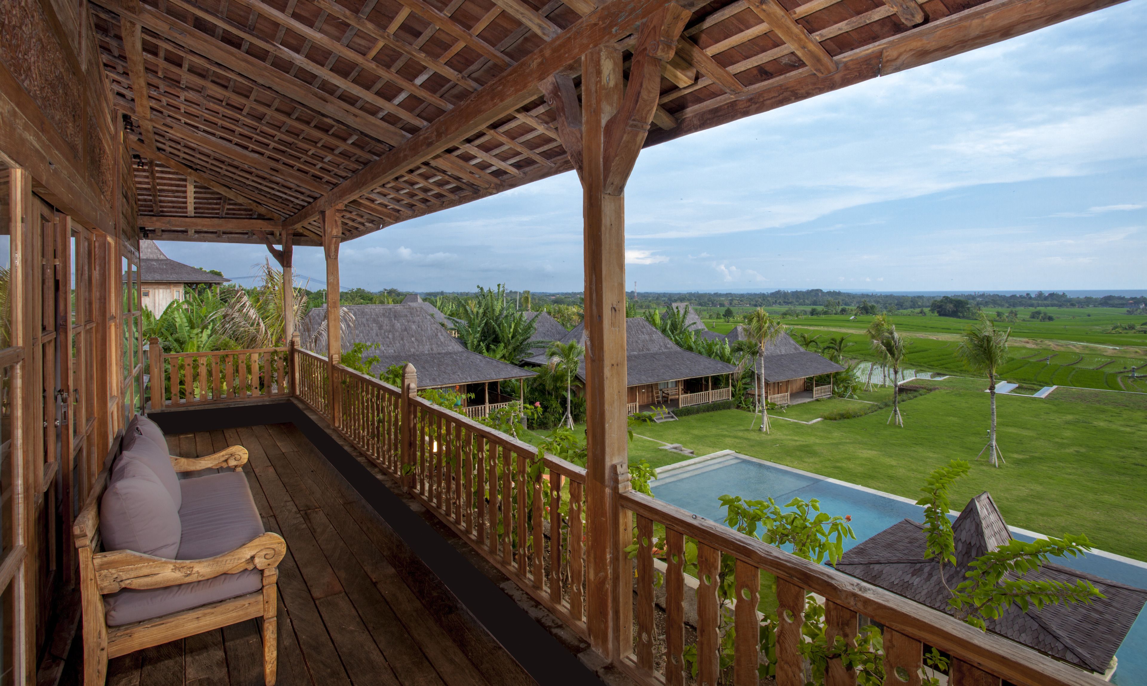 A wooden deck with a bench, overlooking a pool and a lush landscape with thatched-roof huts.