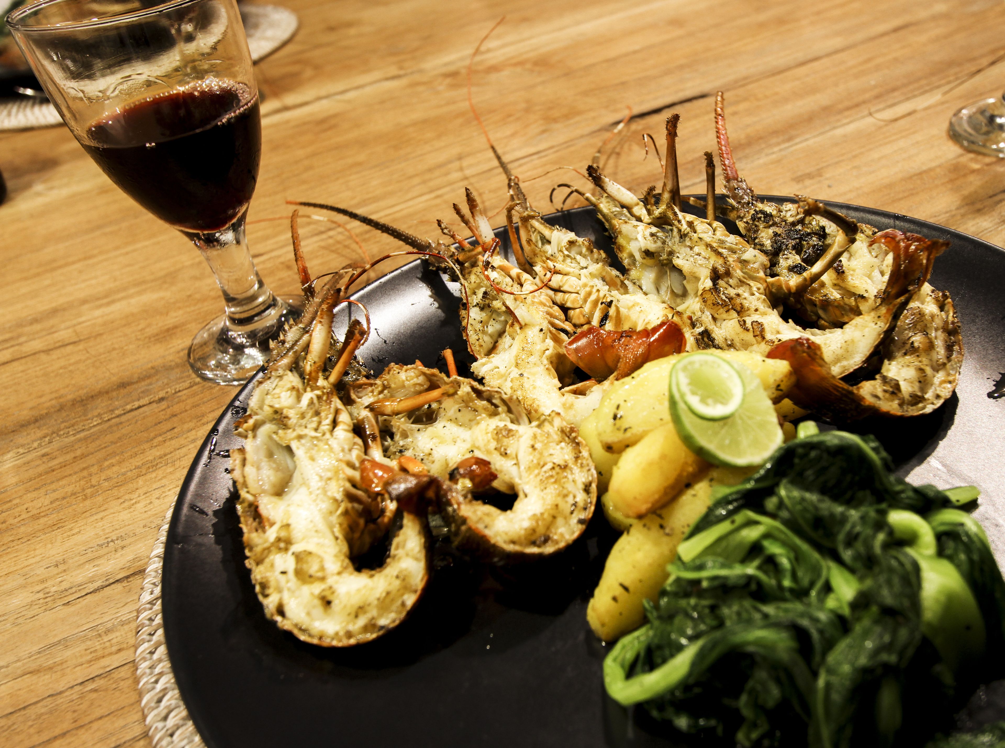 Prato de frutos do mar com lagosta, batatas e vegetais, acompanhado de um copo de vinho tinto sobre uma mesa de madeira.