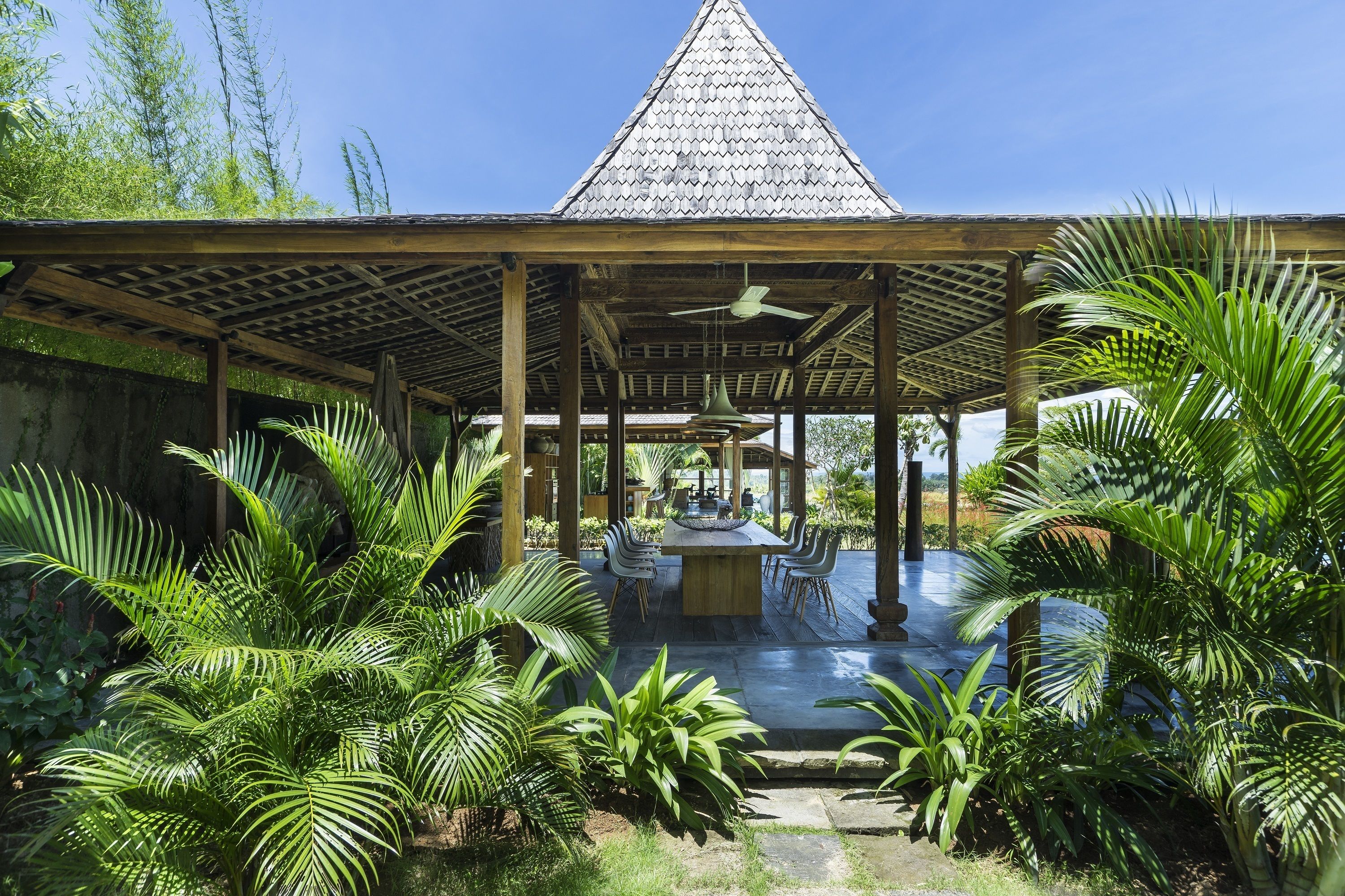 A covered outdoor area with a wooden structure, a ceiling fan, and a central table with chairs.