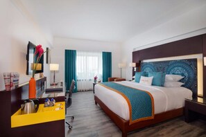 Une chambre d’hôtel avec un grand lit, un bureau avec une chaise, une petite table avec une lampe et une fenêtre avec des rideaux.