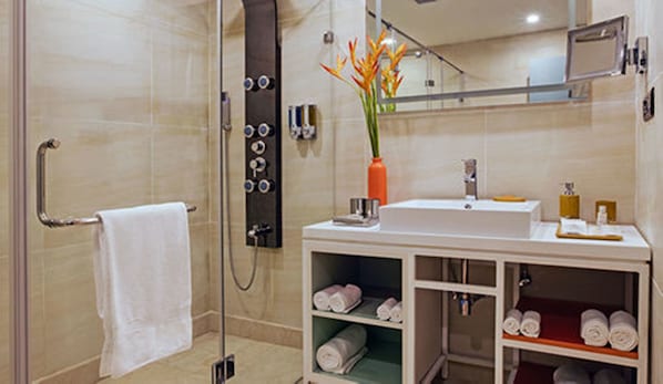A modern bathroom with a glass shower enclosure, a white sink with a mirror above it, and a shelf with neatly rolled towels.