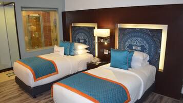 A hotel room with two beds, each with a headboard, a nightstand, a lamp, and a window with blinds.