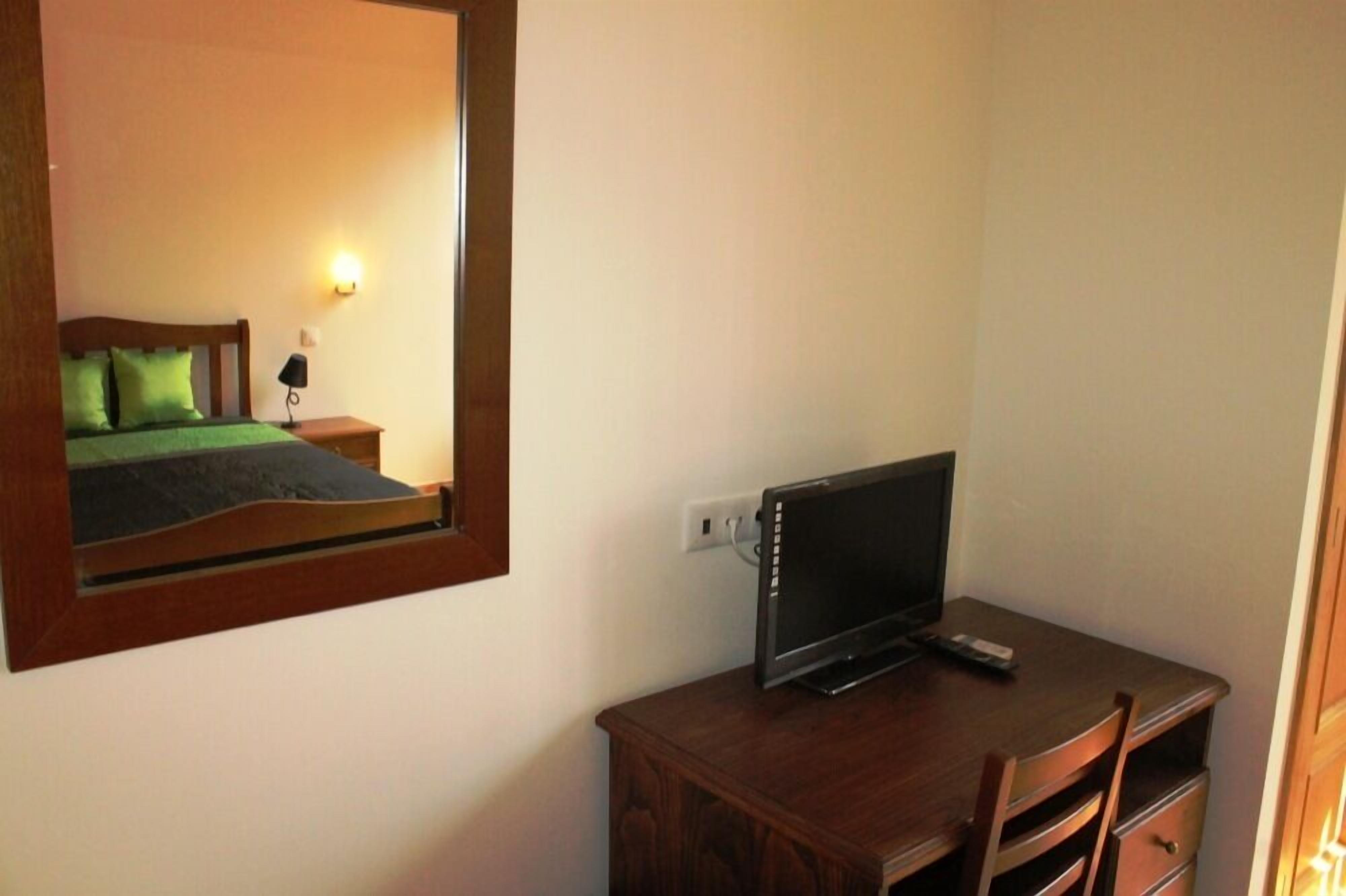 A bedroom with a bed, a bedside table, a TV on a wooden dresser, and a mirror on the wall.