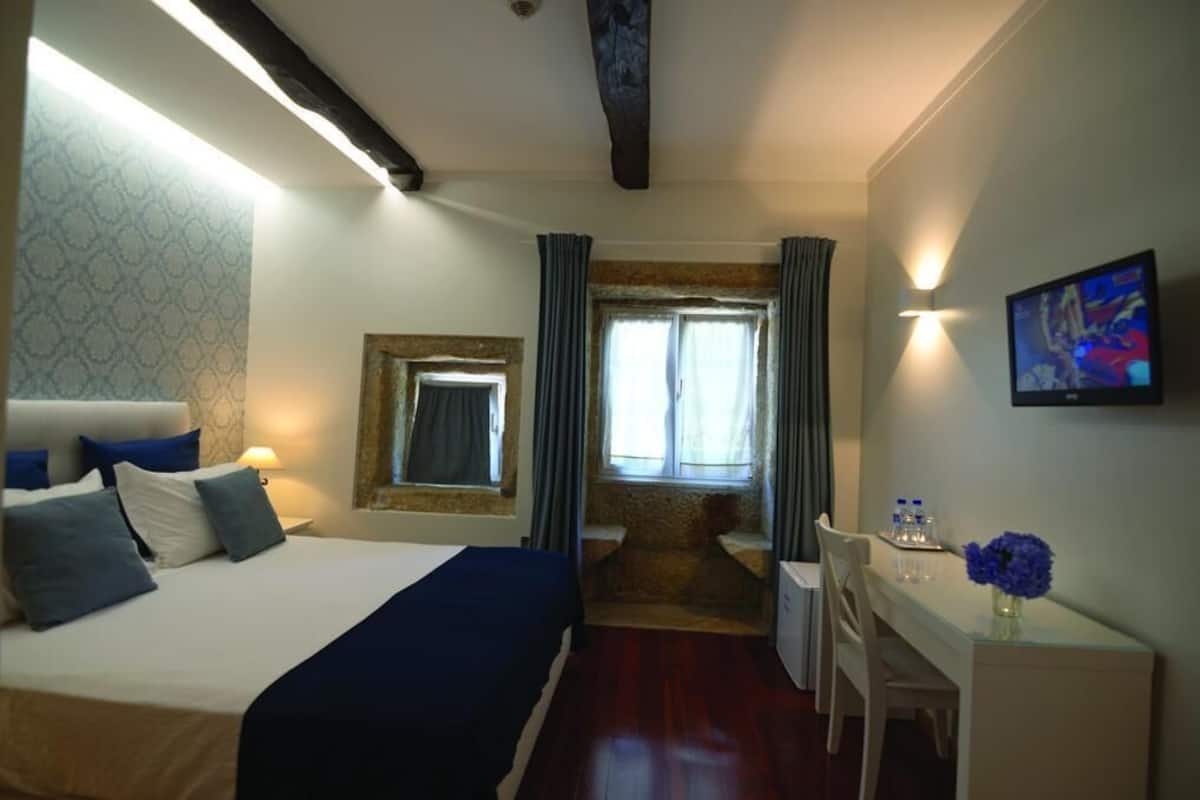 A hotel room with a bed, a desk, a chair, a small table, a TV, and a window with curtains.