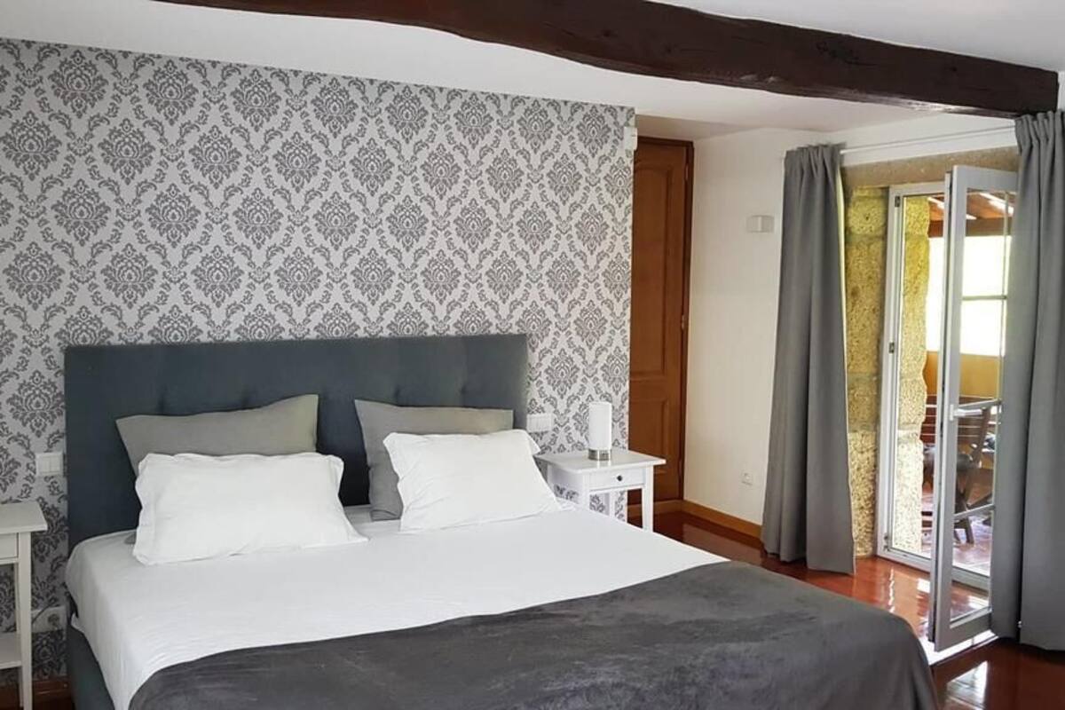 A bedroom with a large bed, a nightstand, a patterned wallpaper, and a view through a glass door.