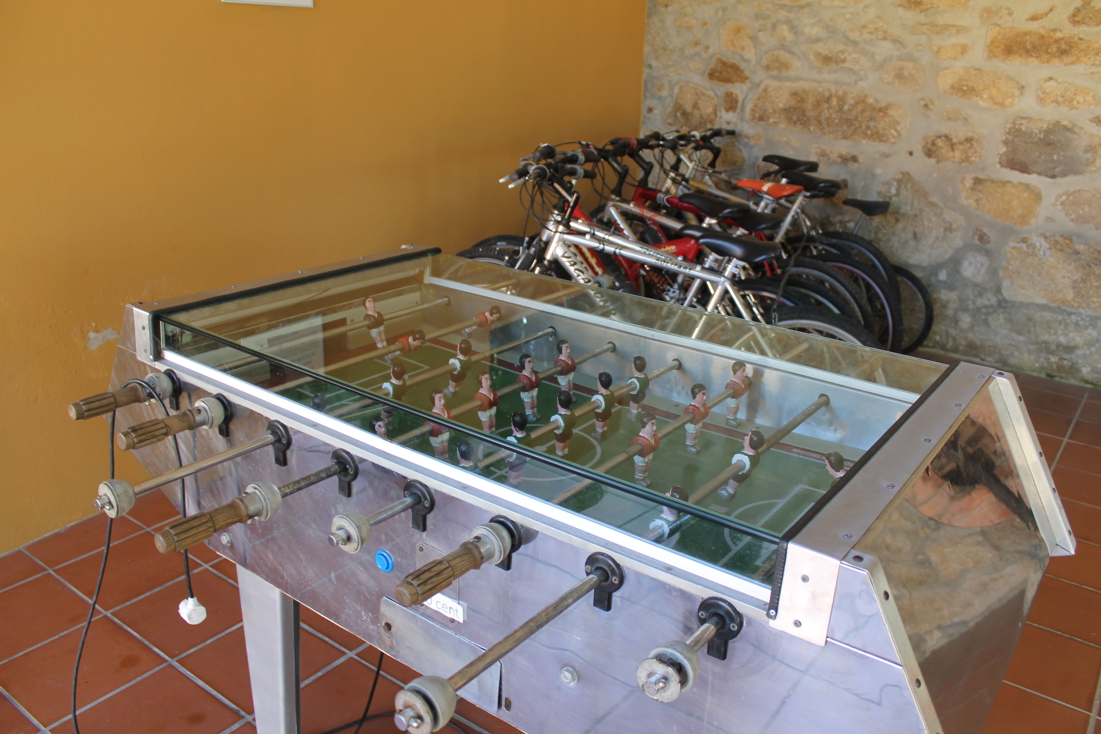 Ein Kicker mit Glasplatte und Metallgestell, platziert in einem Raum mit Steinwand und Fahrrädern im Hintergrund.