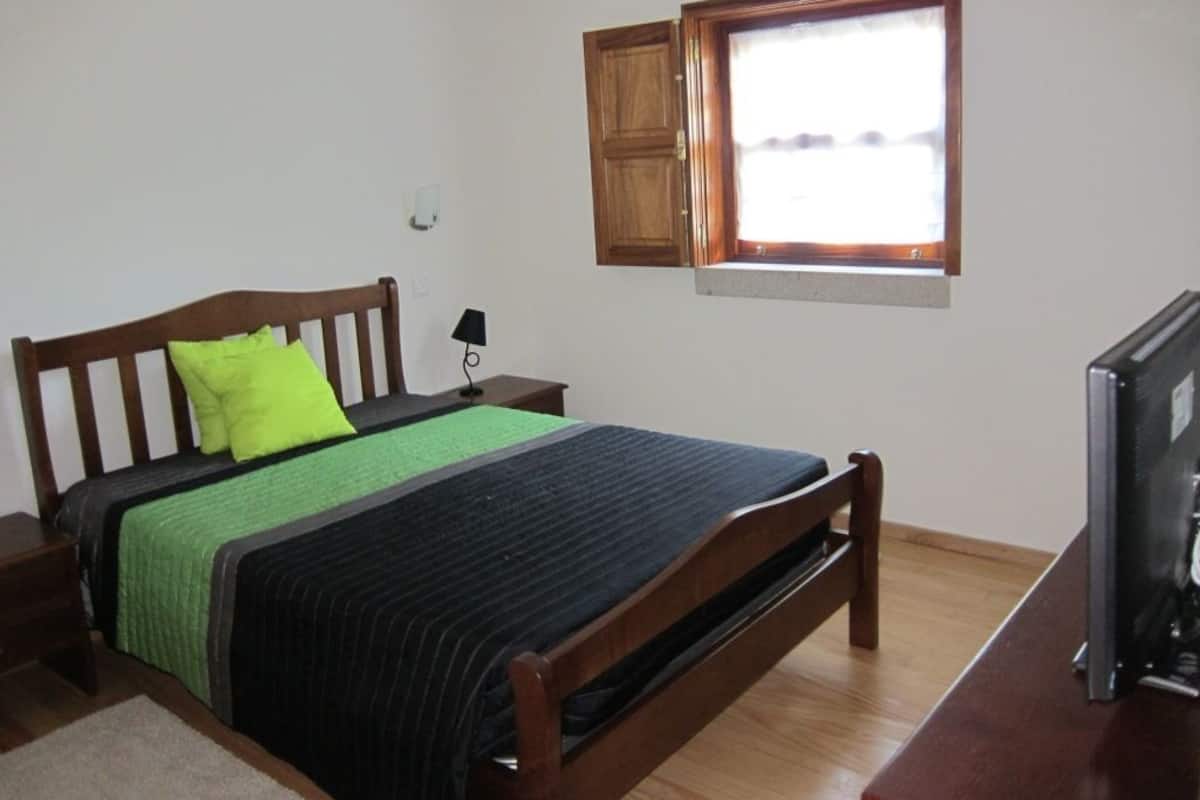 A bedroom with a wooden bed, a green pillow, a black and green bedspread, a small bedside table with a lamp, a wooden cabinet, and a television on a wooden stand.