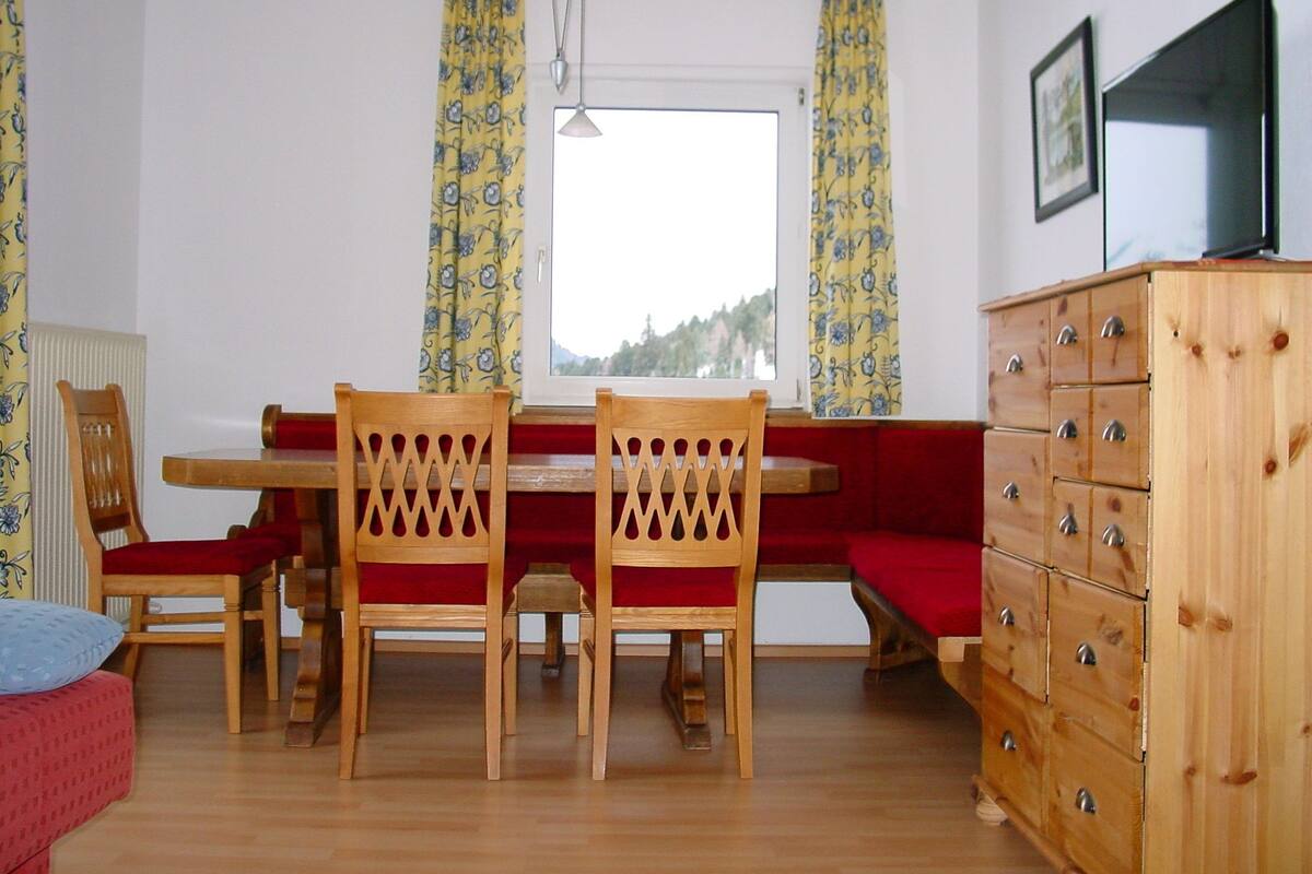 A room with a wooden dining table, two wooden chairs, a red bench, a wooden dresser, and a window with curtains.
