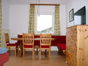 Ein Zimmer mit einem Holztisch, zwei Stühlen, einer roten Bank, einem Holzschrank und einem Fenster mit Vorhängen.