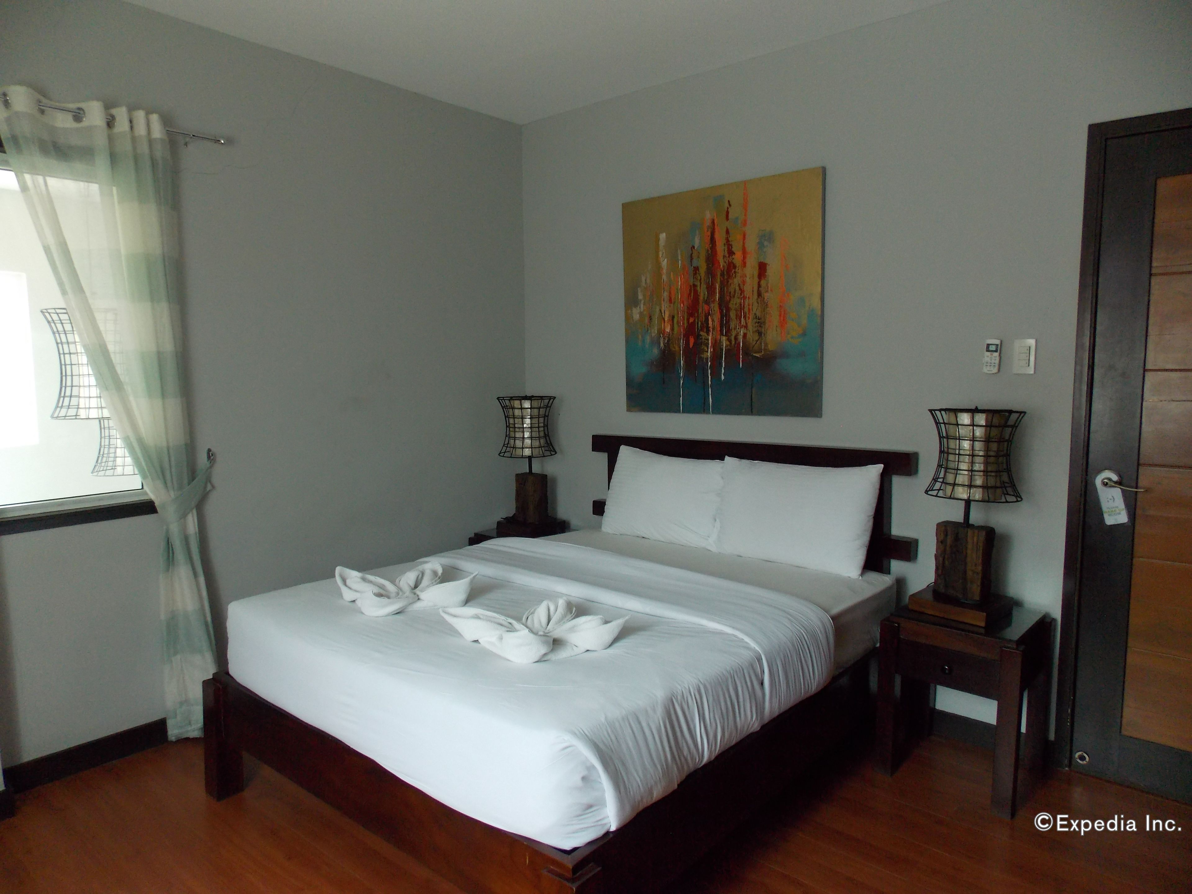A neatly made bed with white linens and towels arranged in a bow shape, a painting on the wall, two bedside lamps, and a wooden floor.