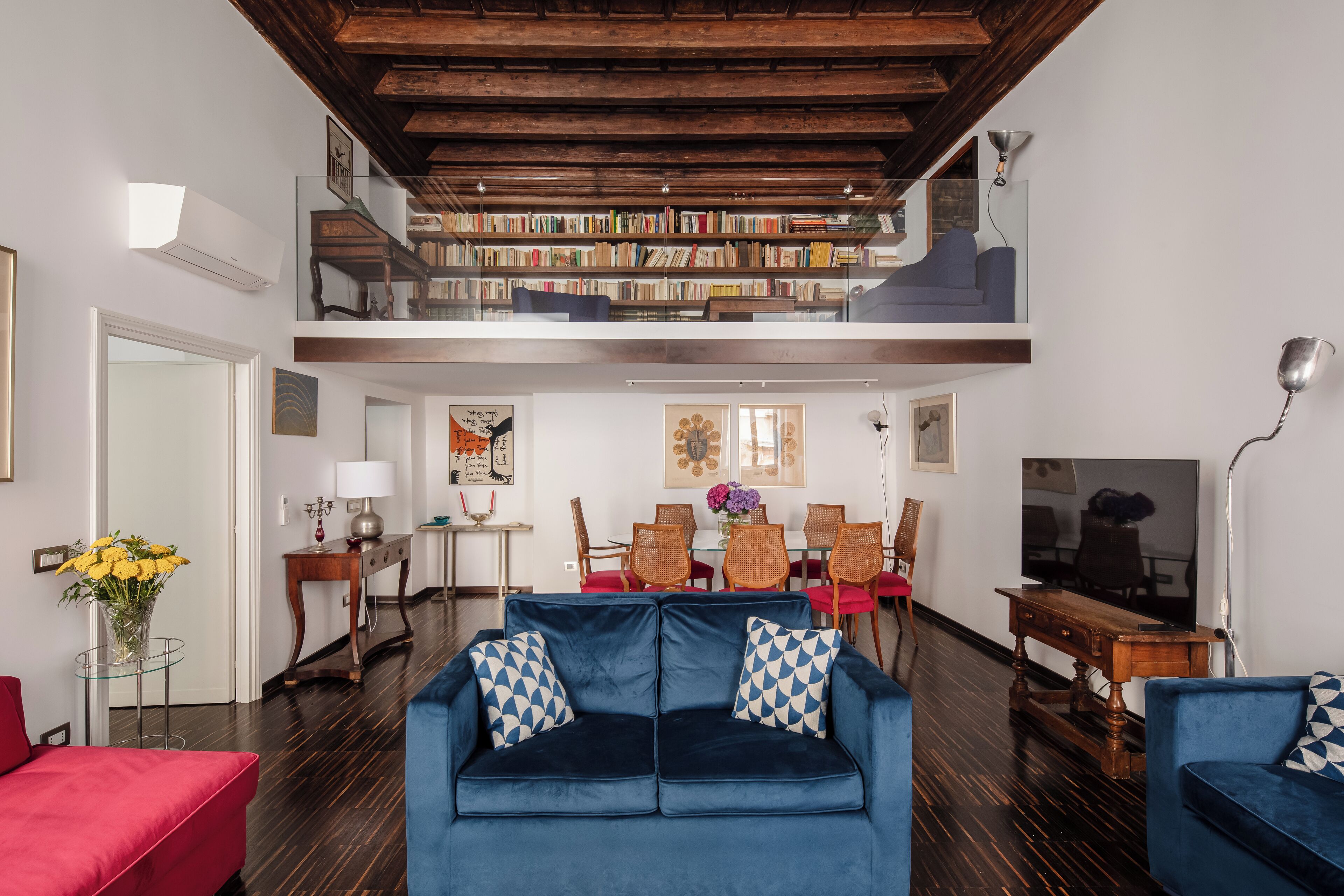 A spacious living room with a blue sofa, a red armchair, and a dining area with a table and chairs. A bookshelf is visible on the upper level, and a flat-screen TV is mounted on the wall.