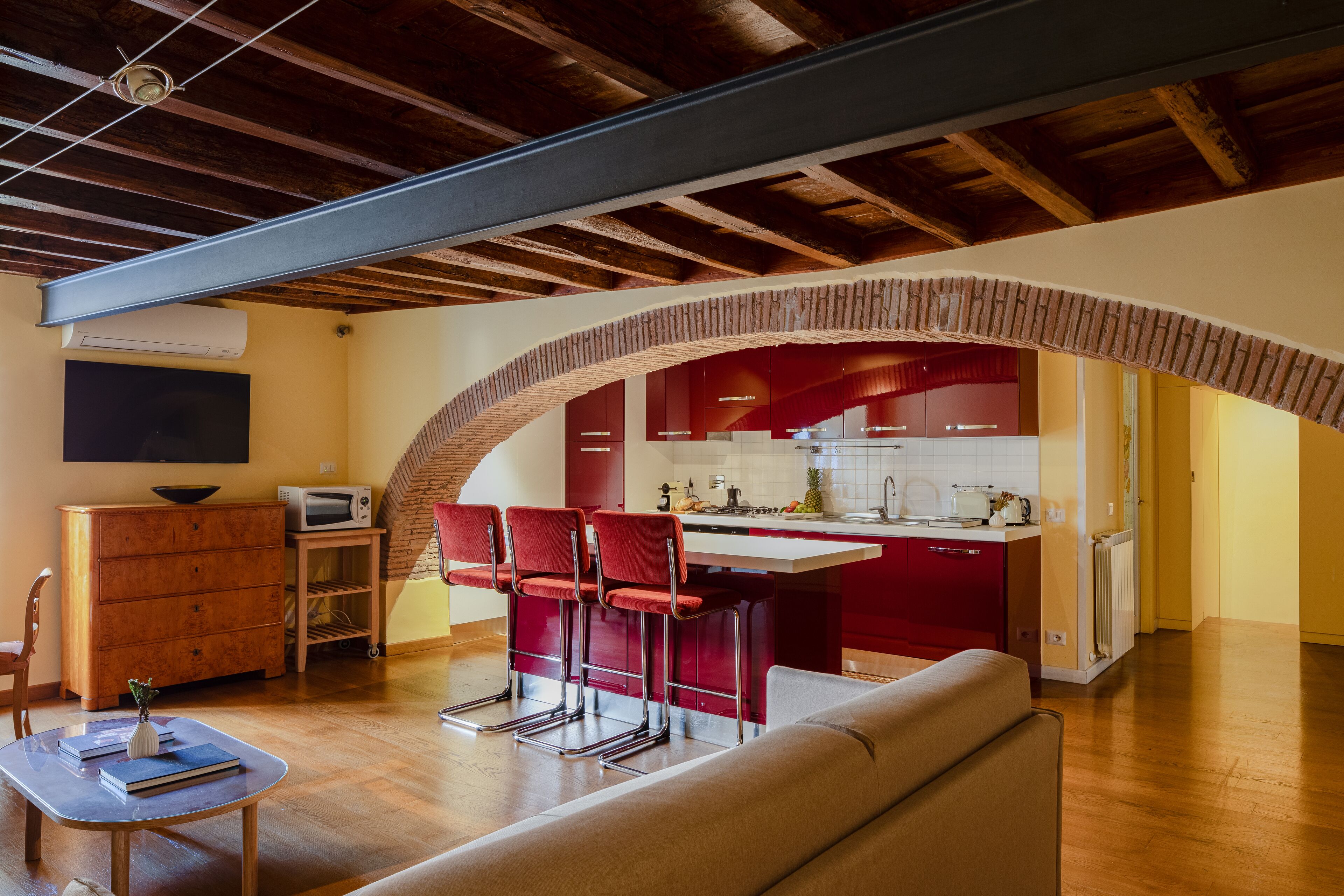 A modern kitchen with red bar stools, a wooden dining table, and a sofa in a living area with exposed wooden beams.