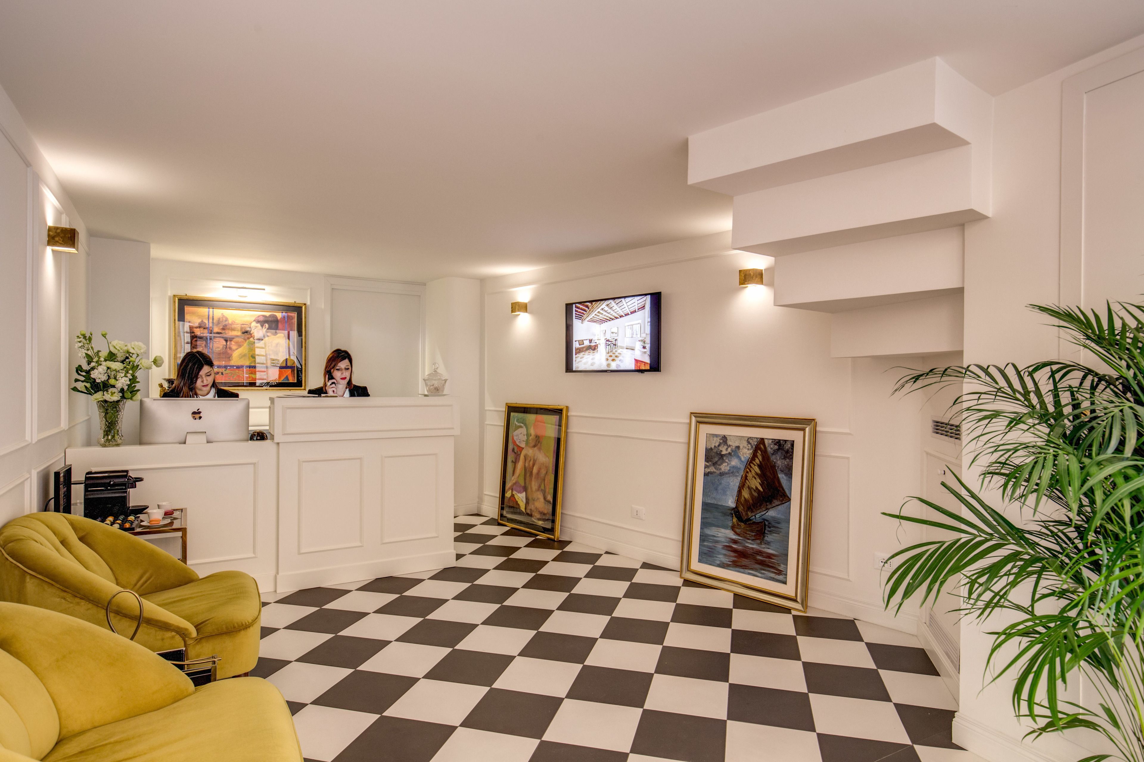 A lobby with a checkered floor, a reception desk, a yellow armchair, framed artwork, and a potted plant.