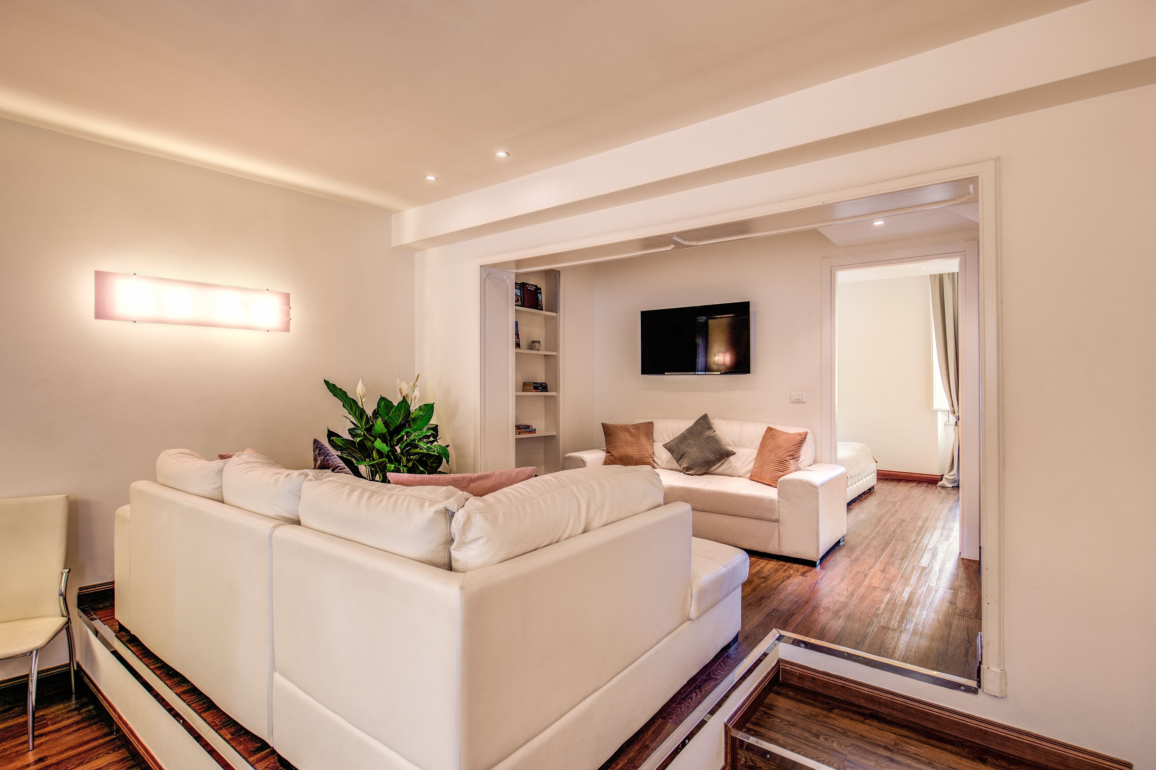 A modern living room with a white sofa, a small table, and a plant. There is a TV mounted on the wall and a door leading to another room.