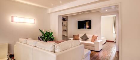 A modern living room with a white sofa, a small table, and a plant. There is a TV mounted on the wall and a door leading to another room.
