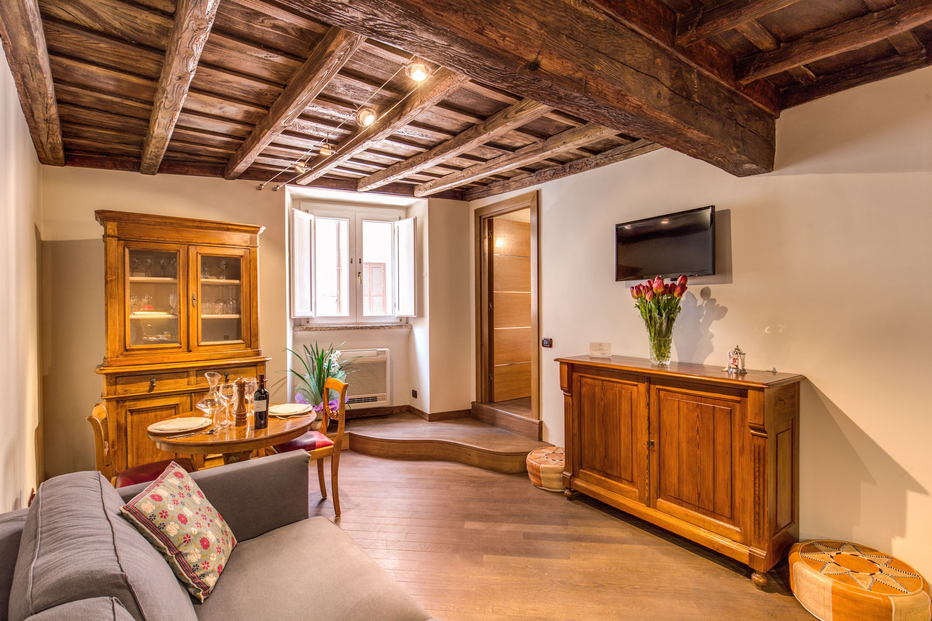 A spacious living area with a wooden ceiling, a dining table with chairs, a sofa, a cabinet, and a television mounted on the wall.