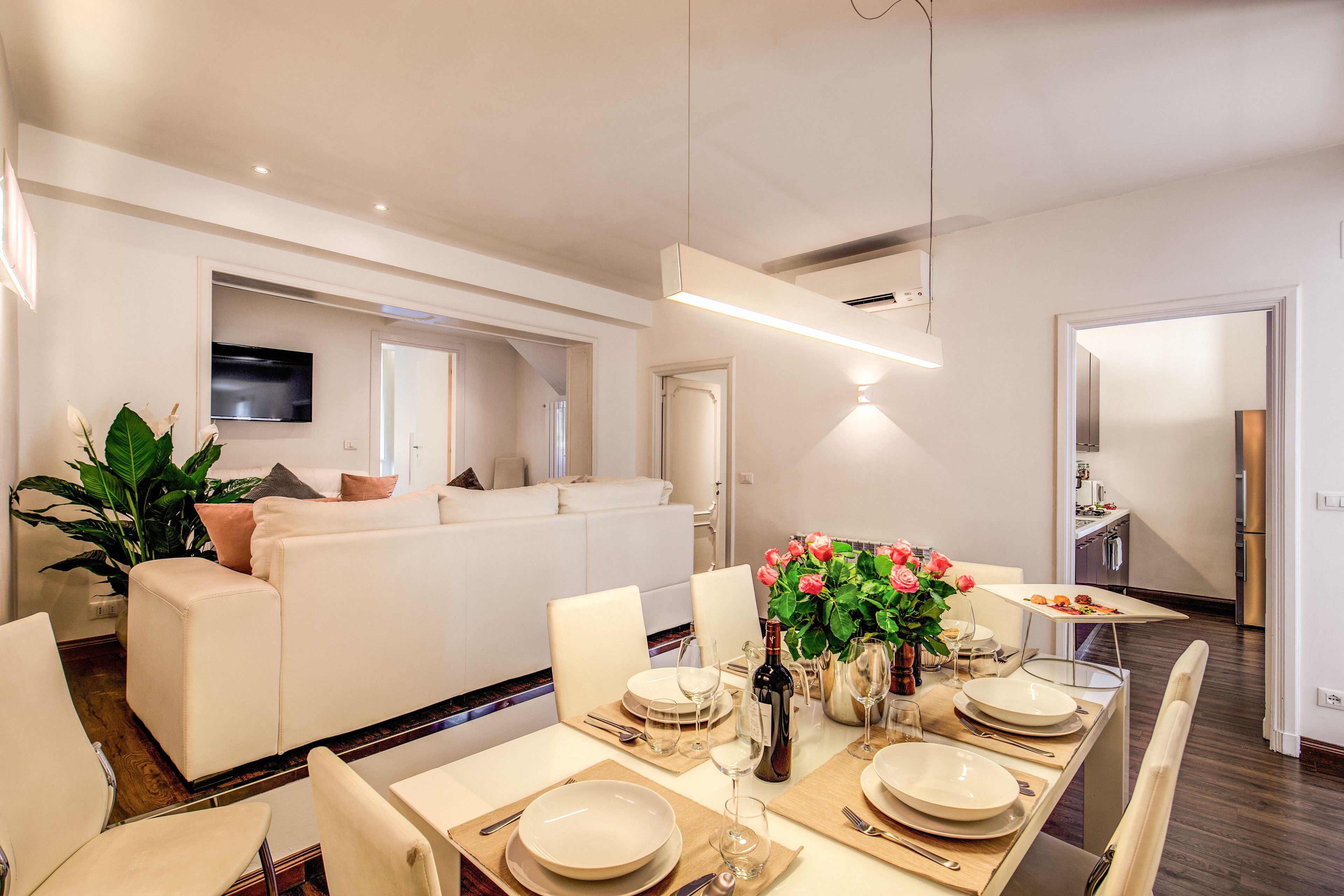A modern dining area with a long table set for six, surrounded by white chairs. A television is mounted on the wall, and a kitchen is visible in the background.