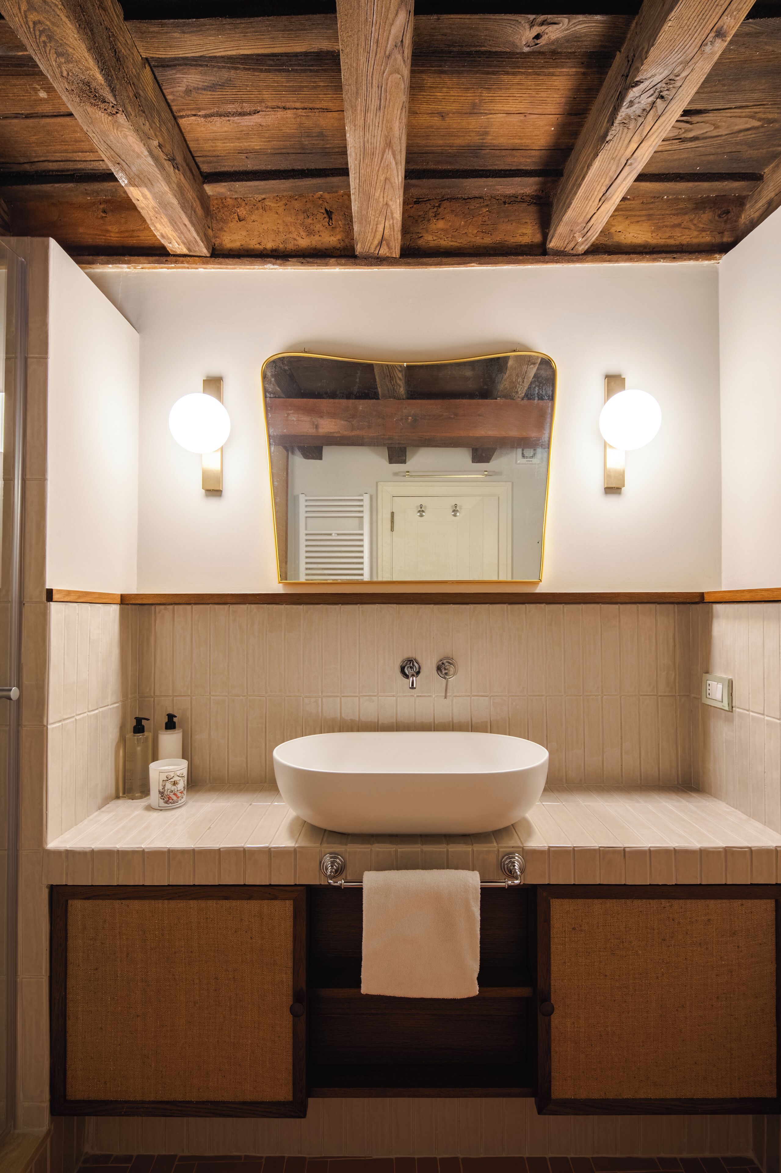 A bathroom with a wooden ceiling, a large mirror, two wall-mounted lights, a white sink, and a towel rack.