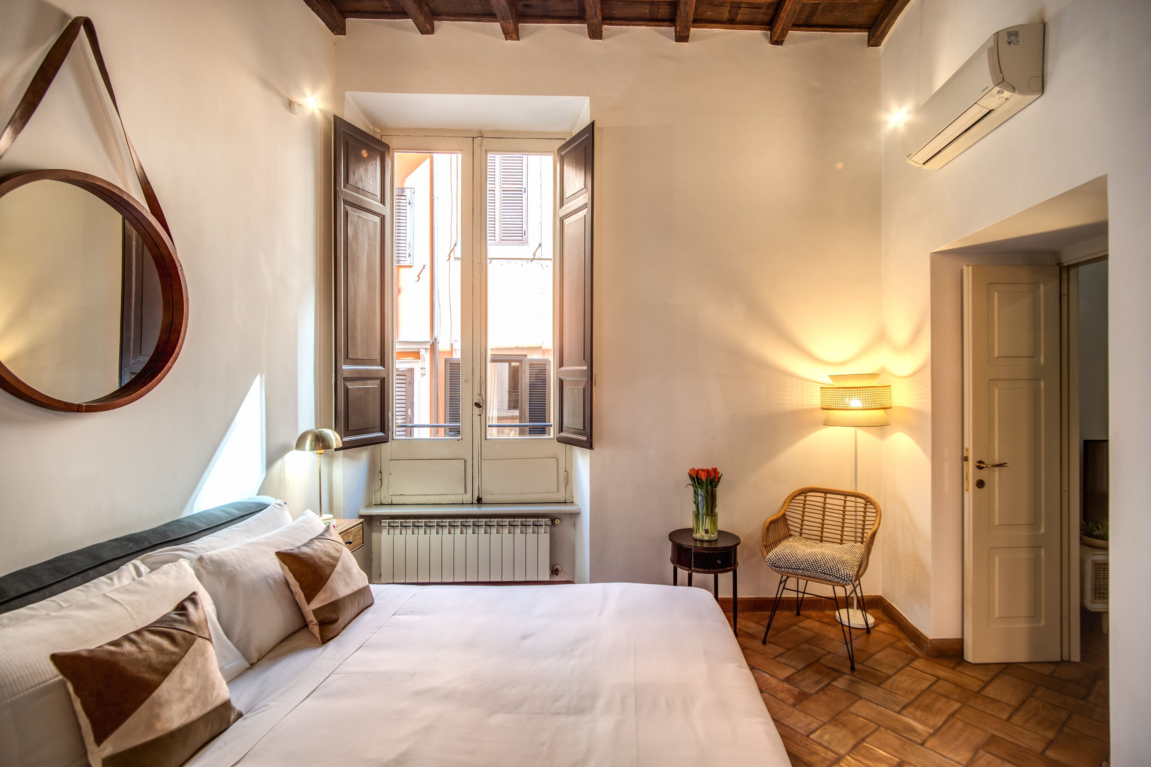 Un dormitorio con una cama grande, una ventana con contraventanas, un espejo montado en la pared, una lámpara de noche, una silla y un equipo de aire acondicionado.