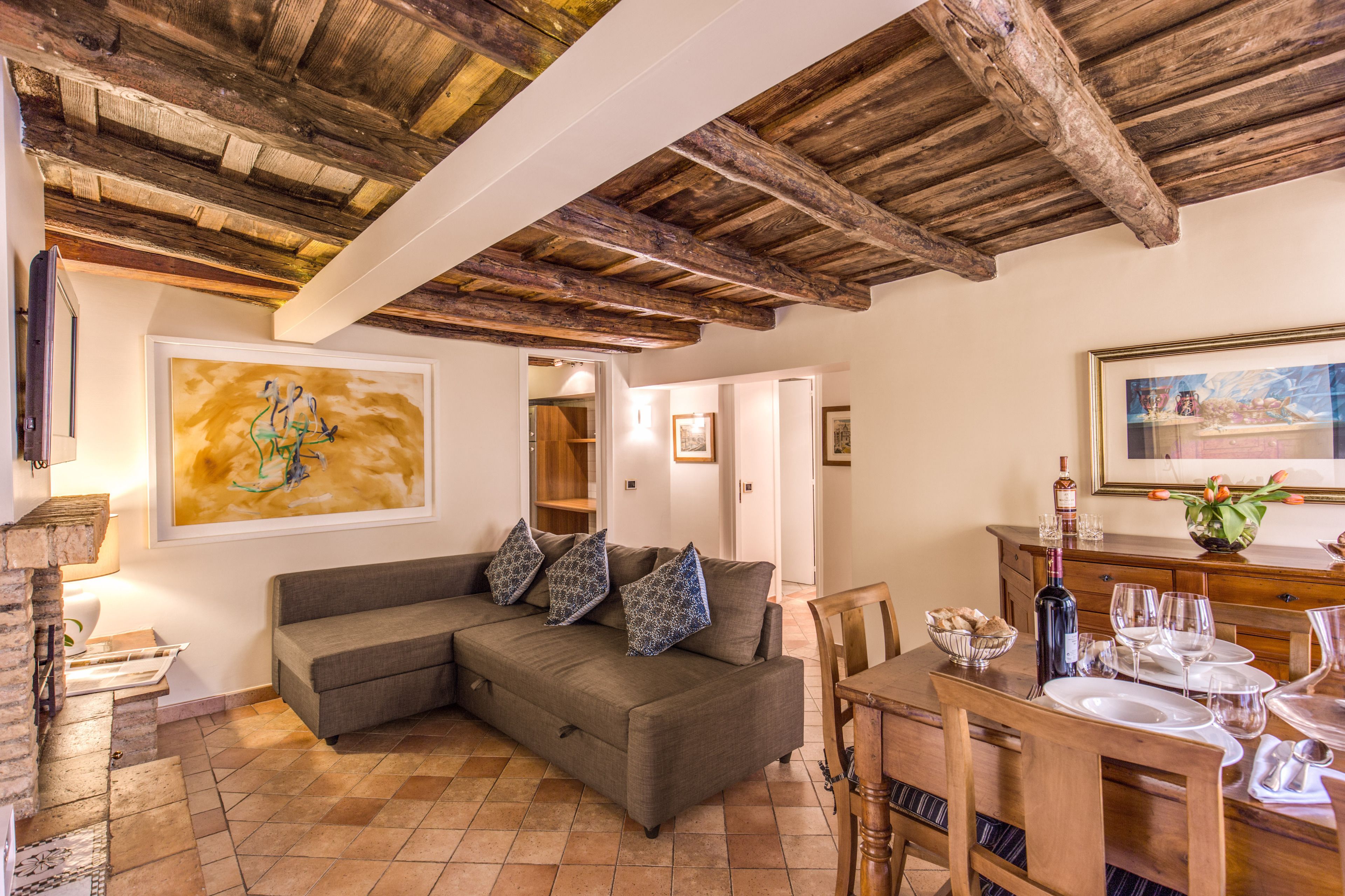 A cozy living room with a wooden ceiling, a gray sectional sofa with patterned cushions, a dining table set for four, and a fireplace.