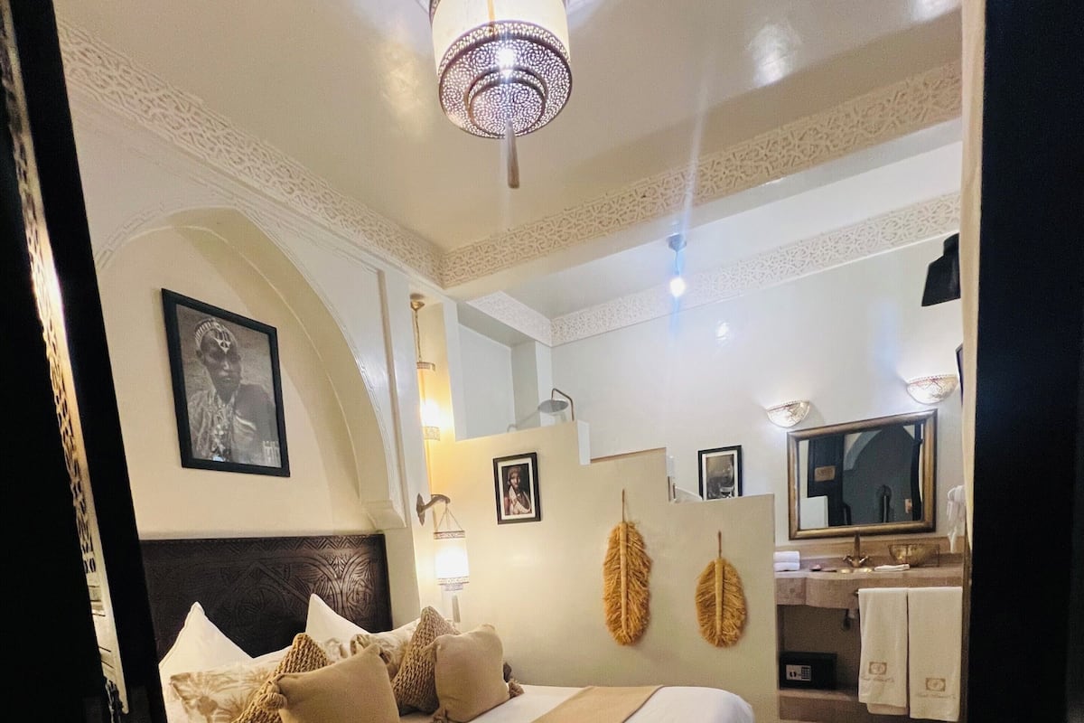 A bedroom with a large bed, a headboard, a chandelier, a mirror, and a bathroom visible in the background.