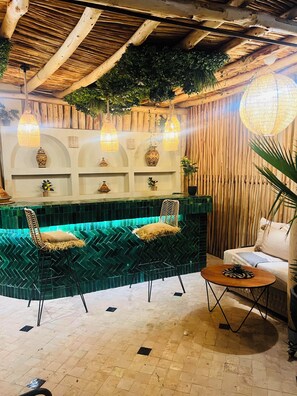 A room with a green patterned sofa, two chairs, a small table, a hanging light fixture, and a wooden wall with shelves. - Riad Reve D'or (Marrakech)