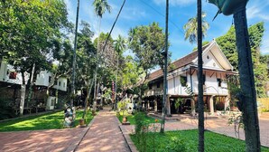 A pathway lined with trees and buildings, leading to a white house with a tiled roof. - Thatsaphone Hotel (Luang Prabang)