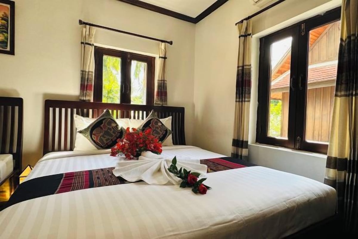 Una cama bien hecha con un ramo de flores rojas, cabecera de madera y una ventana con cortinas.